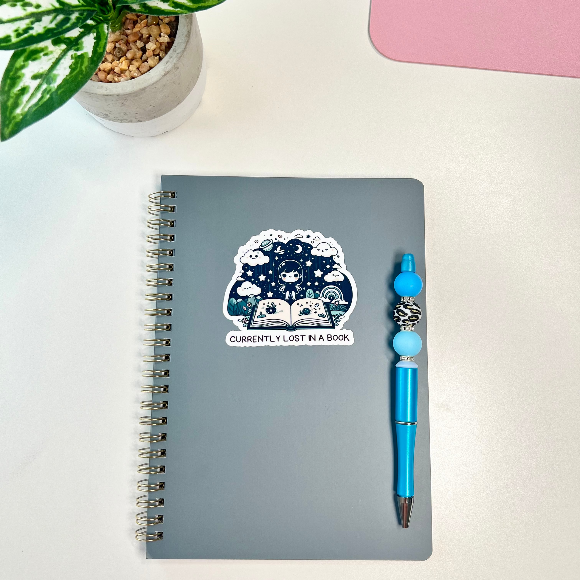 A gray spiral notebook with a sticker that reads 'CURRENTLY LOST IN A BOOK' lies on a white desk next to a blue pen. A potted plant and a pink object are partially visible in the background.