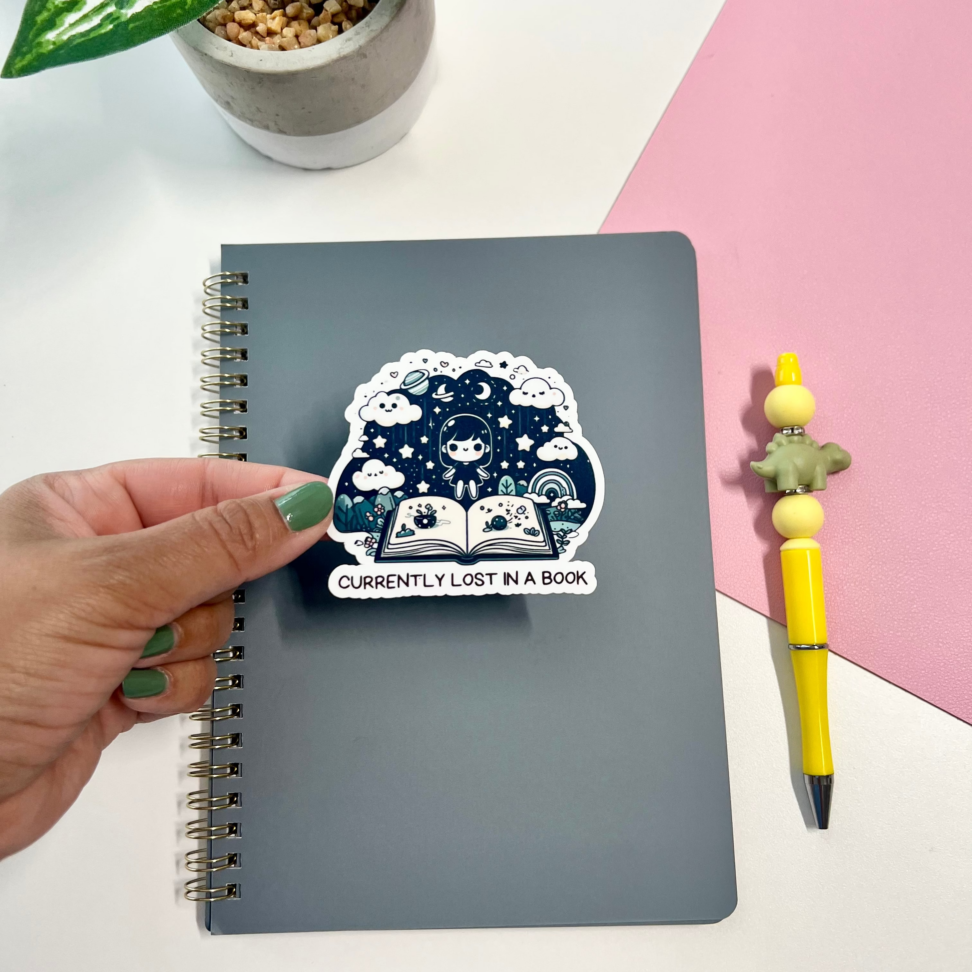 A hand with green nail polish holds a dark gray spiral notebook featuring a sticker that reads 'CURRENTLY LOST IN A BOOK' beneath an illustration of a cozy book under a starry night sky. The scene is styled with a yellow pen on pink paper and a potted plant in the background.