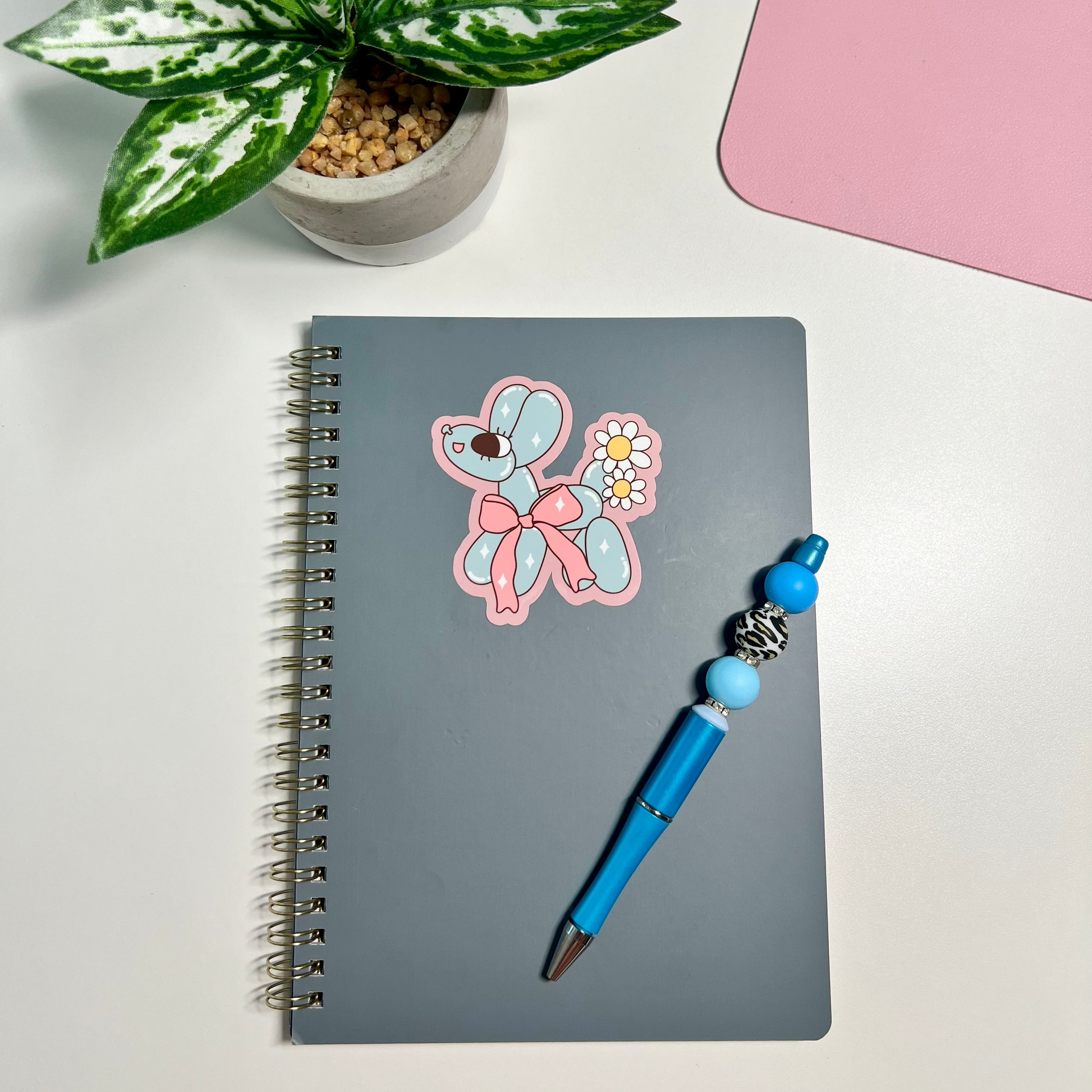 A gray spiral notebook with a pink and blue dog sticker on the cover, paired with a blue pen that has light blue beads and a silver bell, arranged on a white desk next to a potted plant and a pink mat.