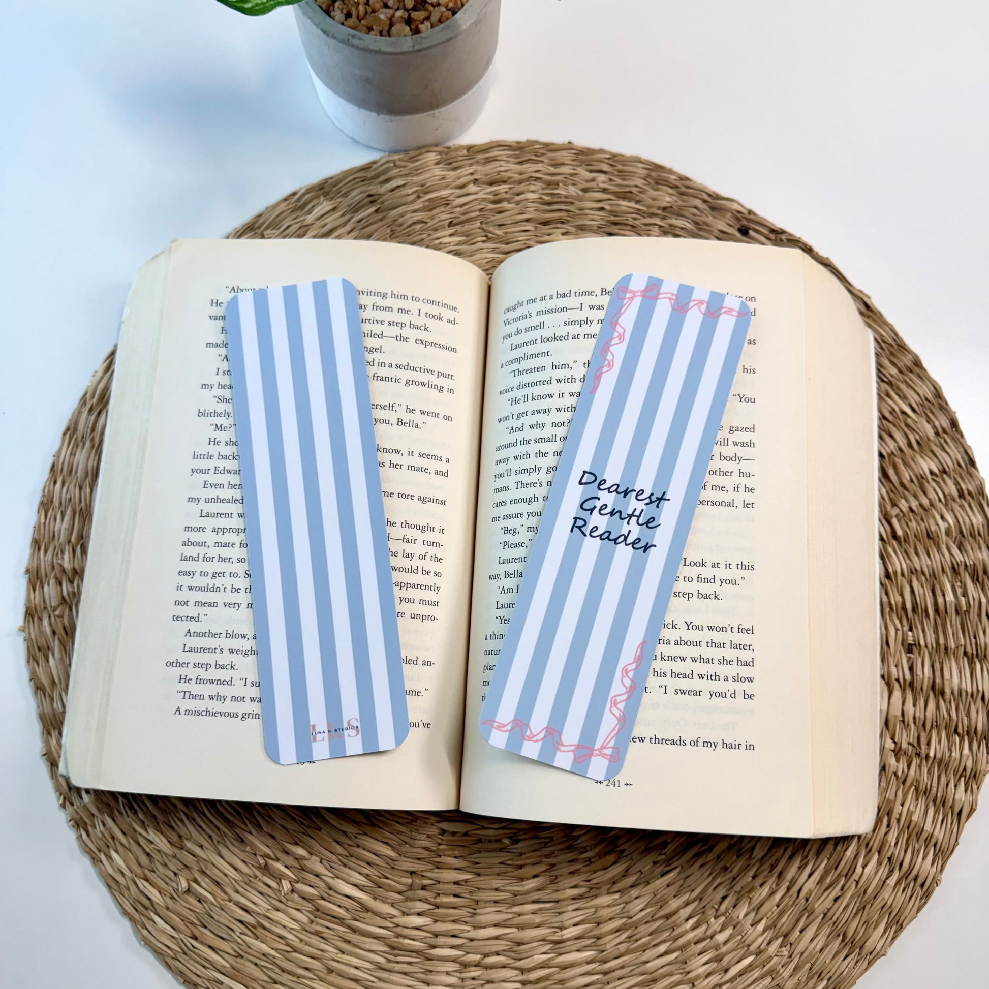 Two decorative paper bookmarks with blue and white stripes are placed on an open book resting on a round woven placemat, with a small potted plant in the background.