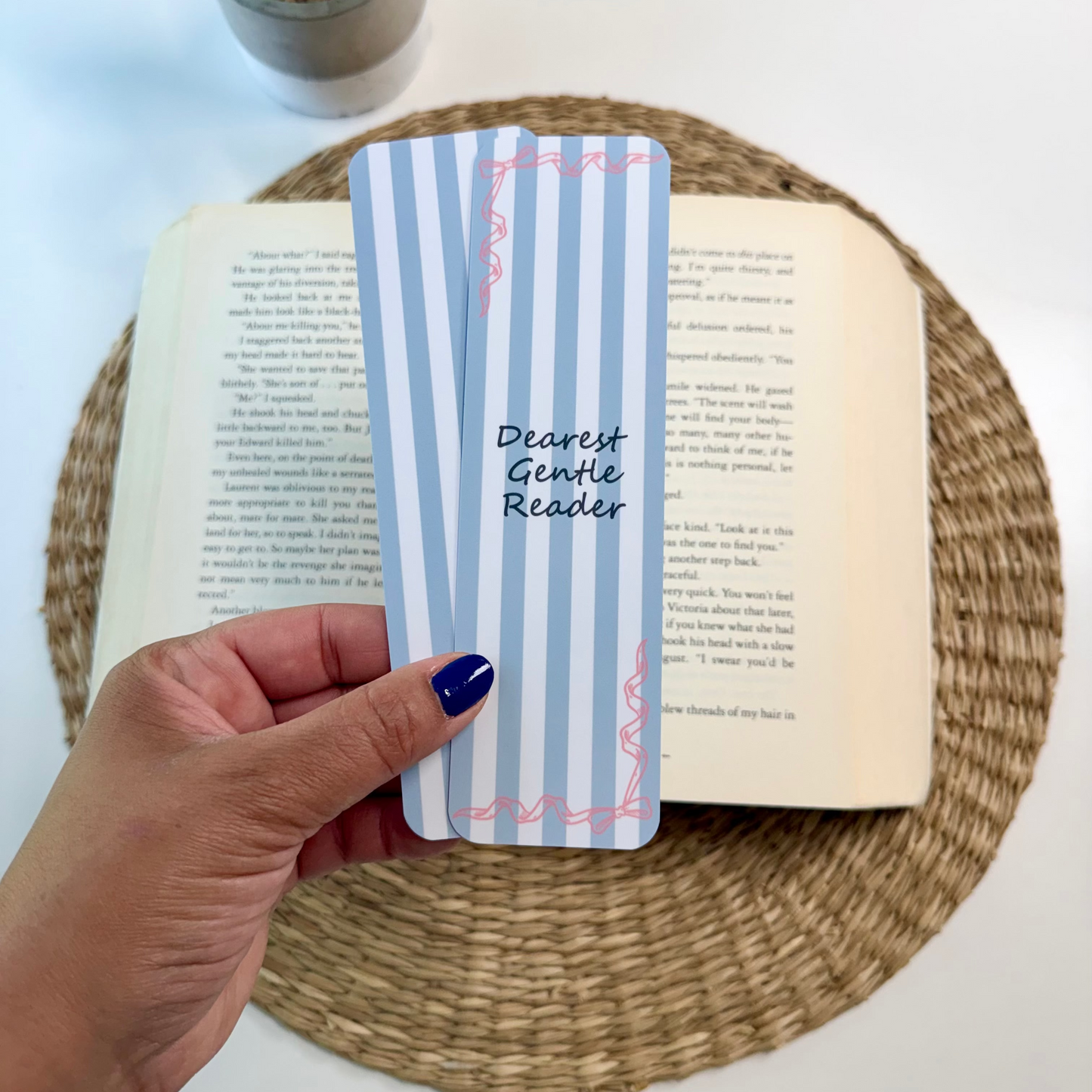 A hand with dark blue nail polish holds a light blue and white striped book bookmark with pink decorative borders and the text 'Dearest Gentle Reader'. The bookmark is positioned over an open book on a round woven placemat, with a small potted plant in the background on a white surface.