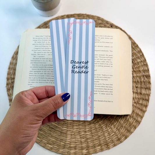 A hand with dark blue nail polish holds a light blue and white striped book bookmark with pink decorative borders and the text 'Dearest Gentle Reader'. The bookmark is positioned over an open book on a round woven placemat, with a small potted plant in the background on a white surface.