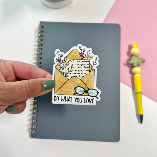 A hand with green nail polish holds a die-cut sticker above a gray spiral notebook. The sticker shows an open envelope with flowers, a letter, and glasses with the text 'DO WHAT YOU LOVE'.