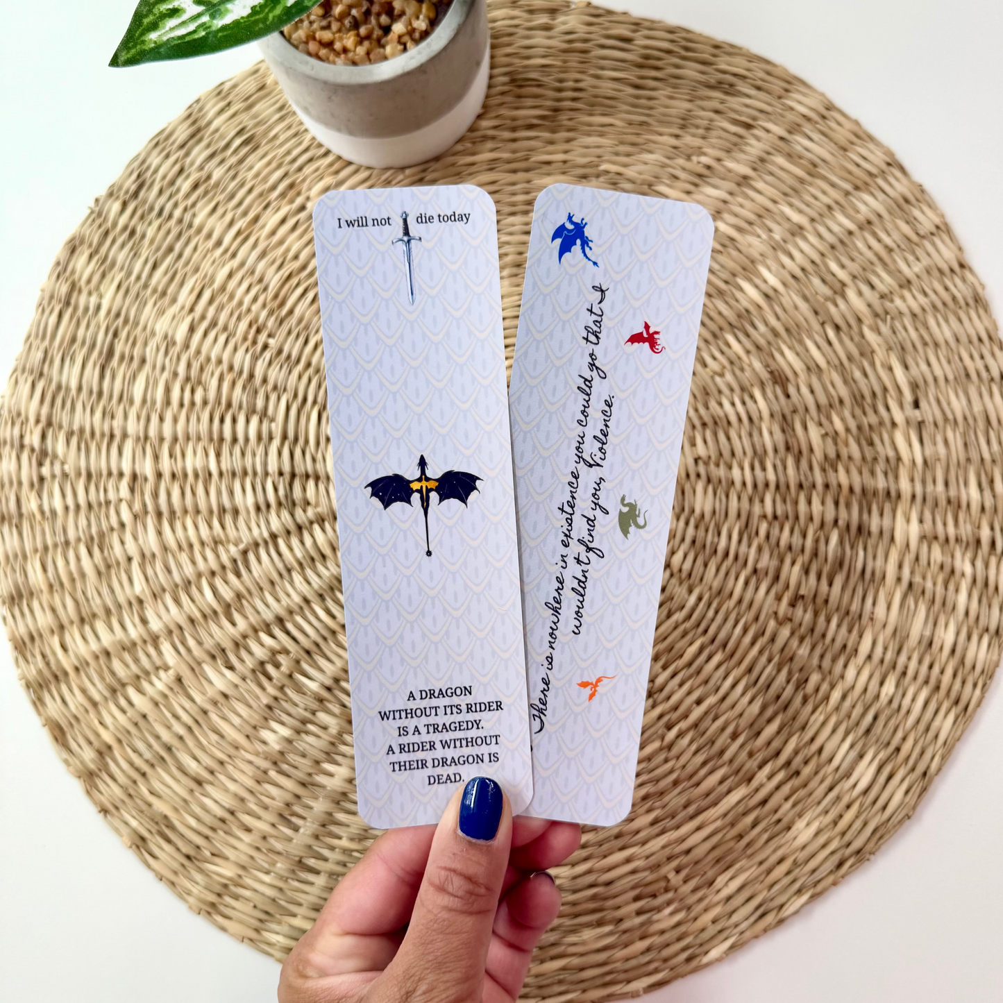 A hand with blue nail polish holds two white bookmarks featuring fantasy illustrations and text, set against a woven round placemat with a potted plant in the background.