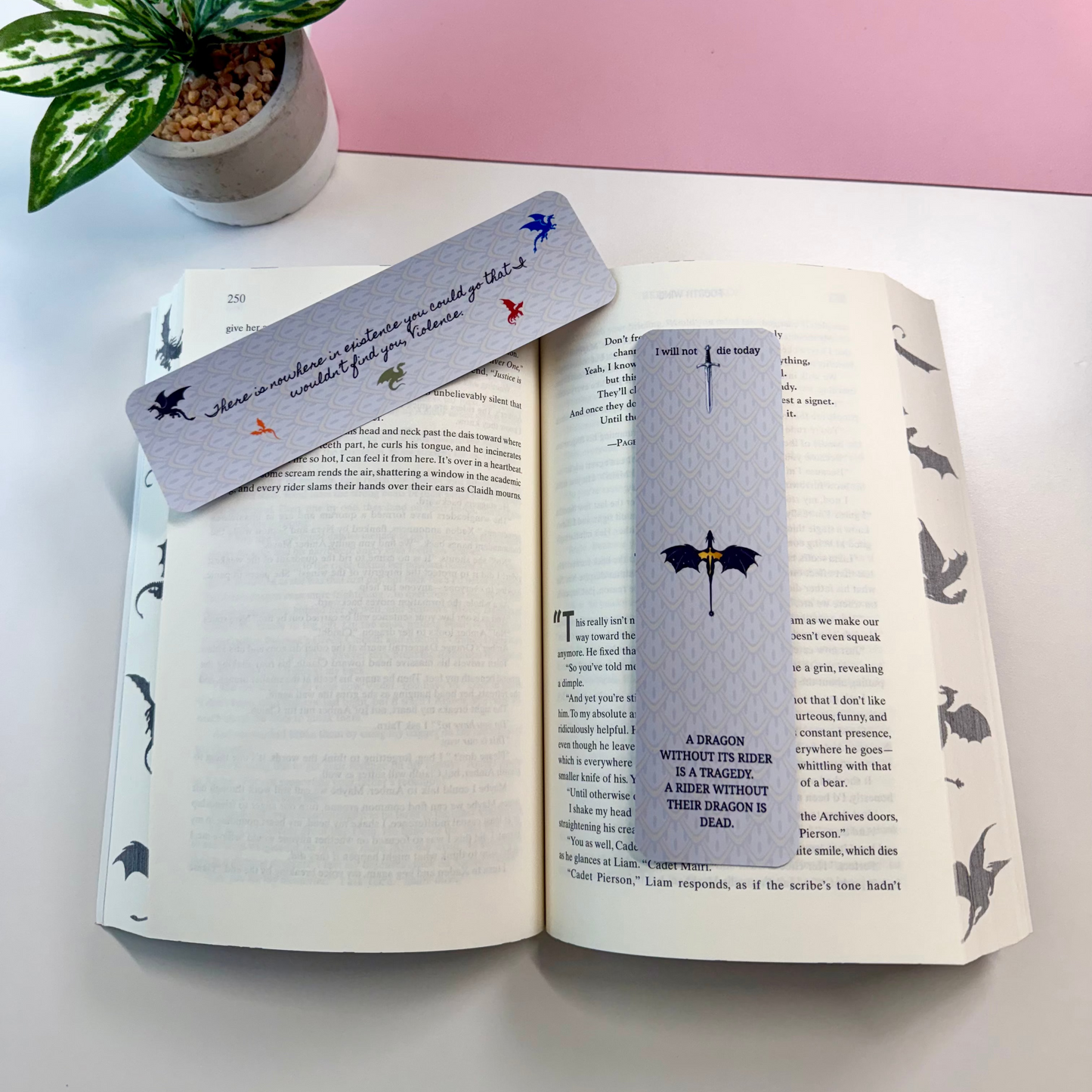 Two decorative book bookmarks with bat and umbrella illustrations are placed on an open book. The scene is styled on a white surface with a pink background panel and a small potted plant in the corner.
