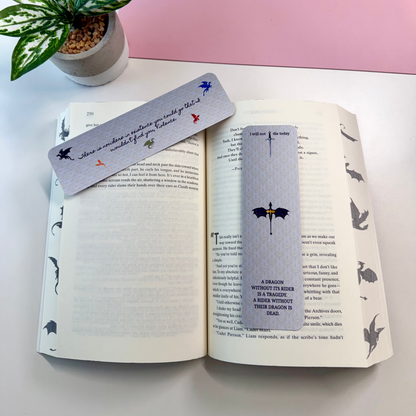 Two decorative book bookmarks with bat and umbrella illustrations are placed on an open book. The scene is styled on a white surface with a pink background panel and a small potted plant in the corner.