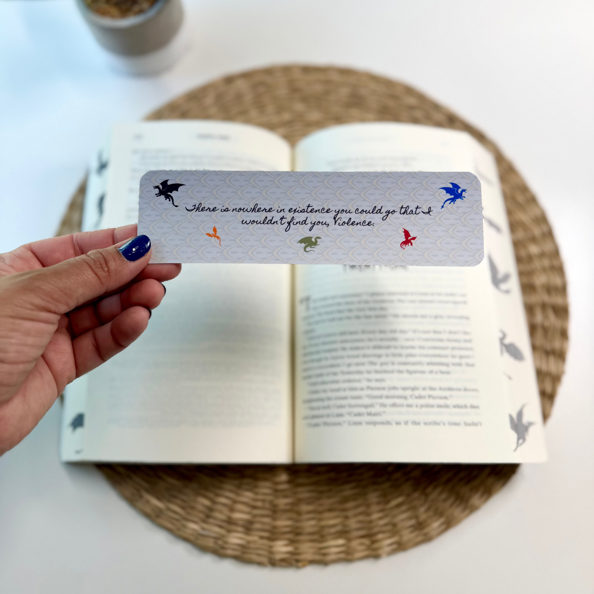 A hand with blue nail polish holds a silver bookmark featuring a quote and small colorful dragon illustrations over an open book on a woven placemat, with a potted plant in the background.