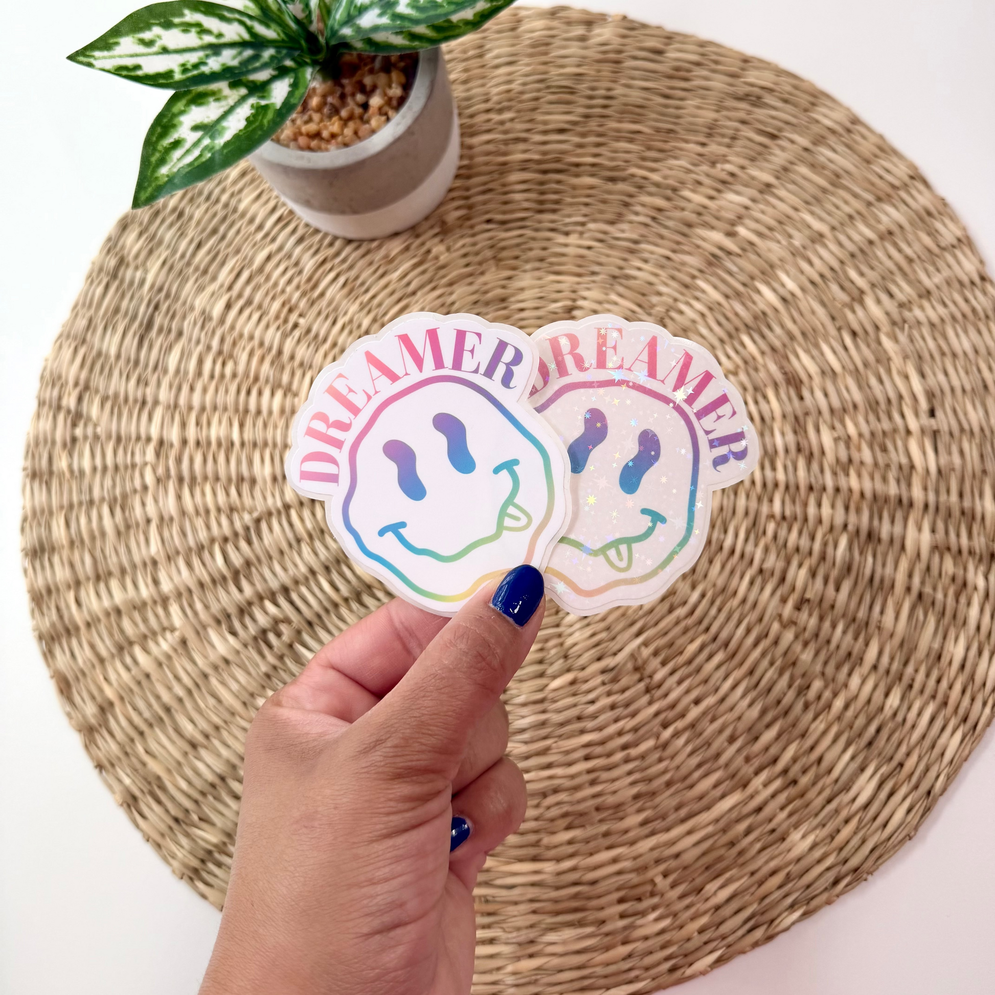 Two die-cut stickers featuring a rainbow smiley face and the word 'DREAMER' in pink and blue lettering, held by a hand over a woven placemat with a small potted plant in the background.