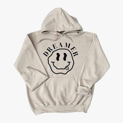 A light beige pullover hoodie with a black graphic on the chest featuring a smiley face with one eye and the word 'DREAMER' arched above it, displayed against a light gray background.