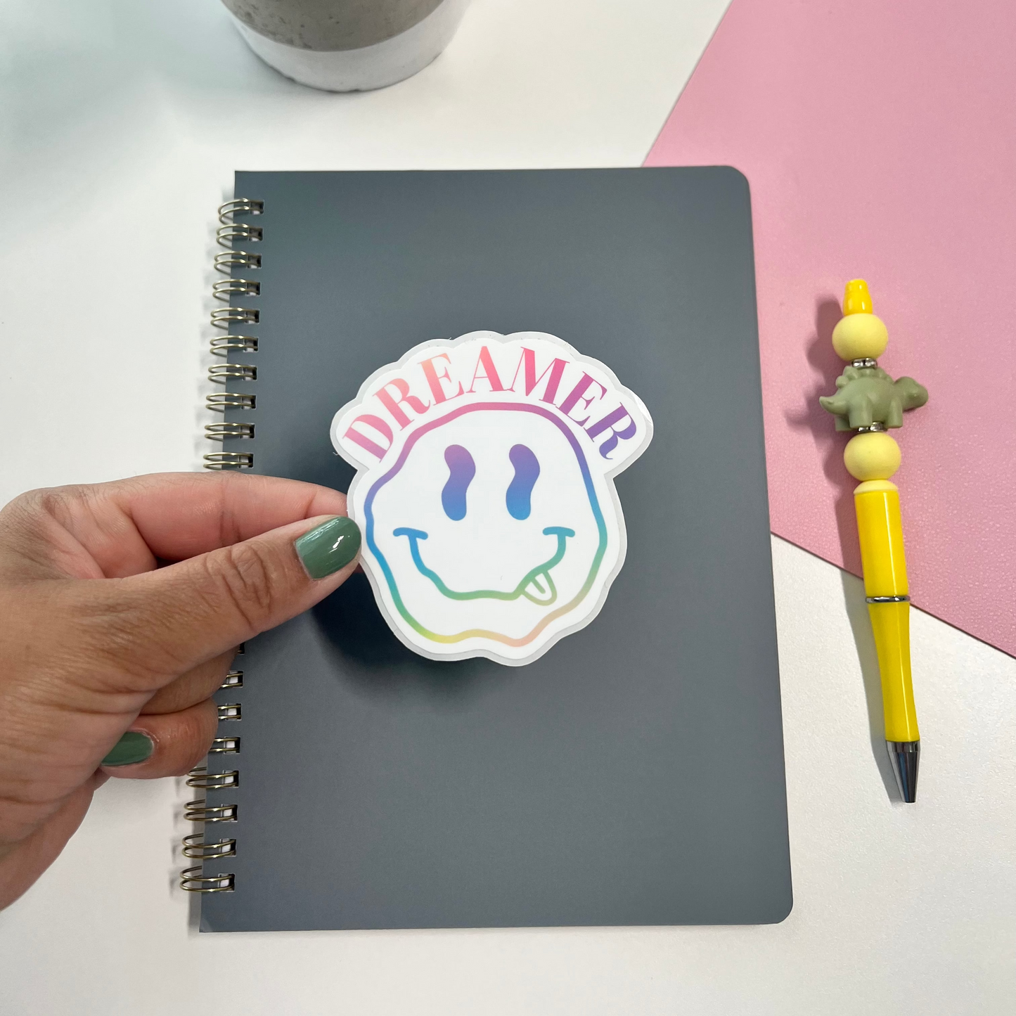Hand holding a sticker with a smiley face and 'DREAMER' text above a notebook on a desk with a plant and pen.
