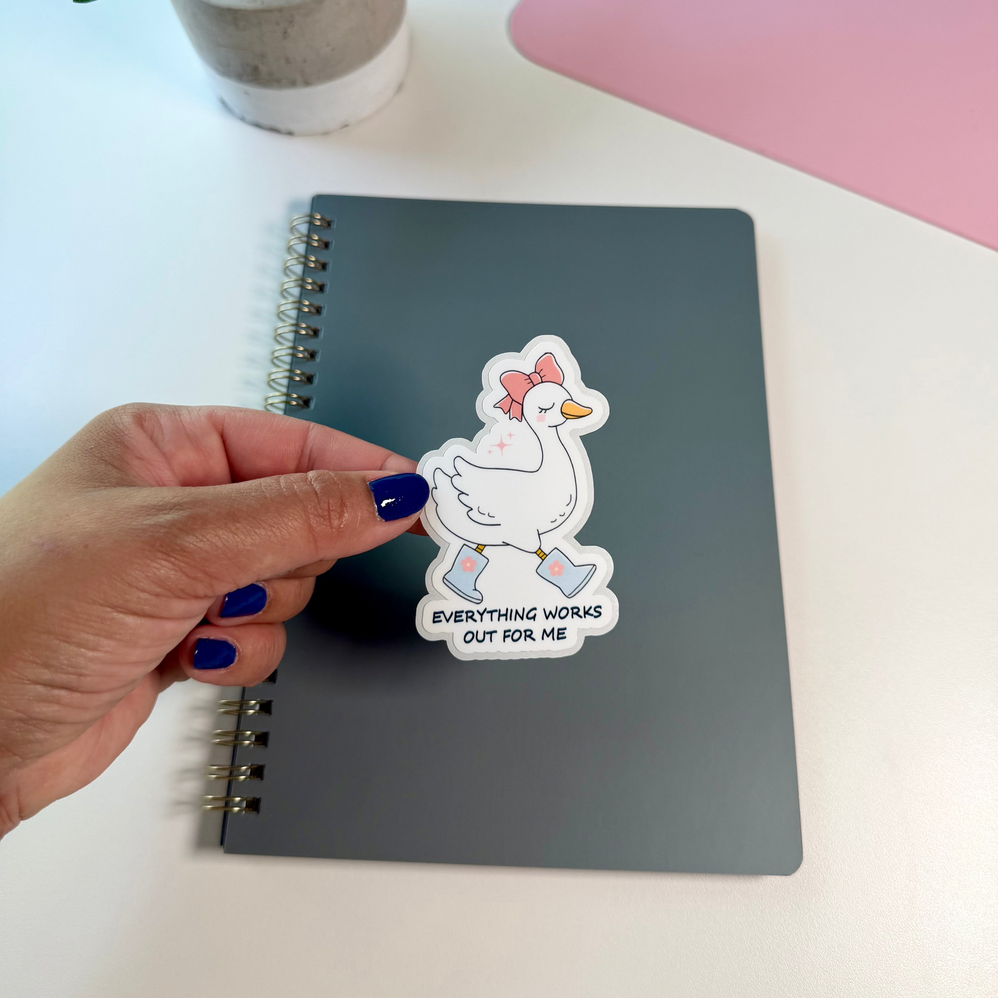 A hand with dark blue nail polish holds a die-cut sticker featuring a white duck with a pink bow and the text 'EVERYTHING WROME OUT FOR ME' above a dark gray spiral notebook on a white desk. A small potted plant and a pink laptop are in the background.