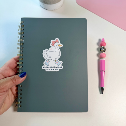 A dark gray spiral notebook with a cartoon duck sticker that reads 'EVERYTHING WORKS OUT FOR ME' is held by a hand on a white desk next to a pink pen, with a small potted plant and pink laptop edge in the background.