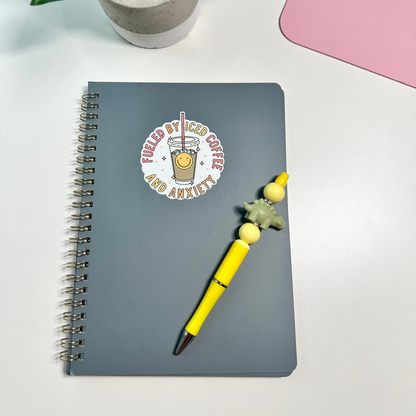 A gray spiral notebook with a circular sticker that reads 'FUELED BY ICED COFFEE AND ANXIETY' in red and white, styled on a white desk next to a yellow pen with a beaded handle and a potted plant.