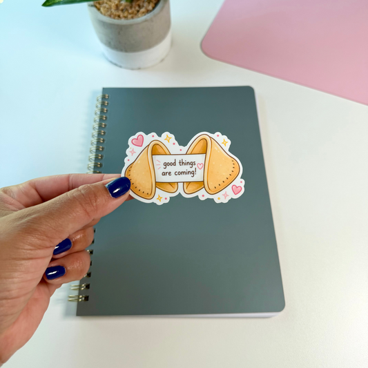 A hand with dark blue nail polish holds a die-cut sticker featuring two yellow fortune cookies with the text 'good things are coming!' and small pink hearts. The sticker is positioned over a gray spiral notebook on a white desk, with a potted plant and a pink desk mat visible in the background.