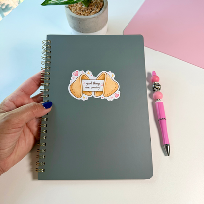 A dark gray spiral notebook with a cute cookie sticker on the cover, held by a hand with blue nail polish on a white desk next to a pink pen.
