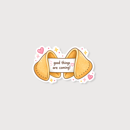 A cute cartoon fortune cookie sticker with the text 'good things are coming!' on a white banner, surrounded by pink hearts, yellow stars, and small pink dots on a light gray background.