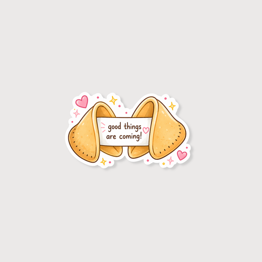 A cute cartoon fortune cookie sticker with the text 'good things are coming!' on a white banner, surrounded by pink hearts, yellow stars, and small pink dots on a light gray background.
