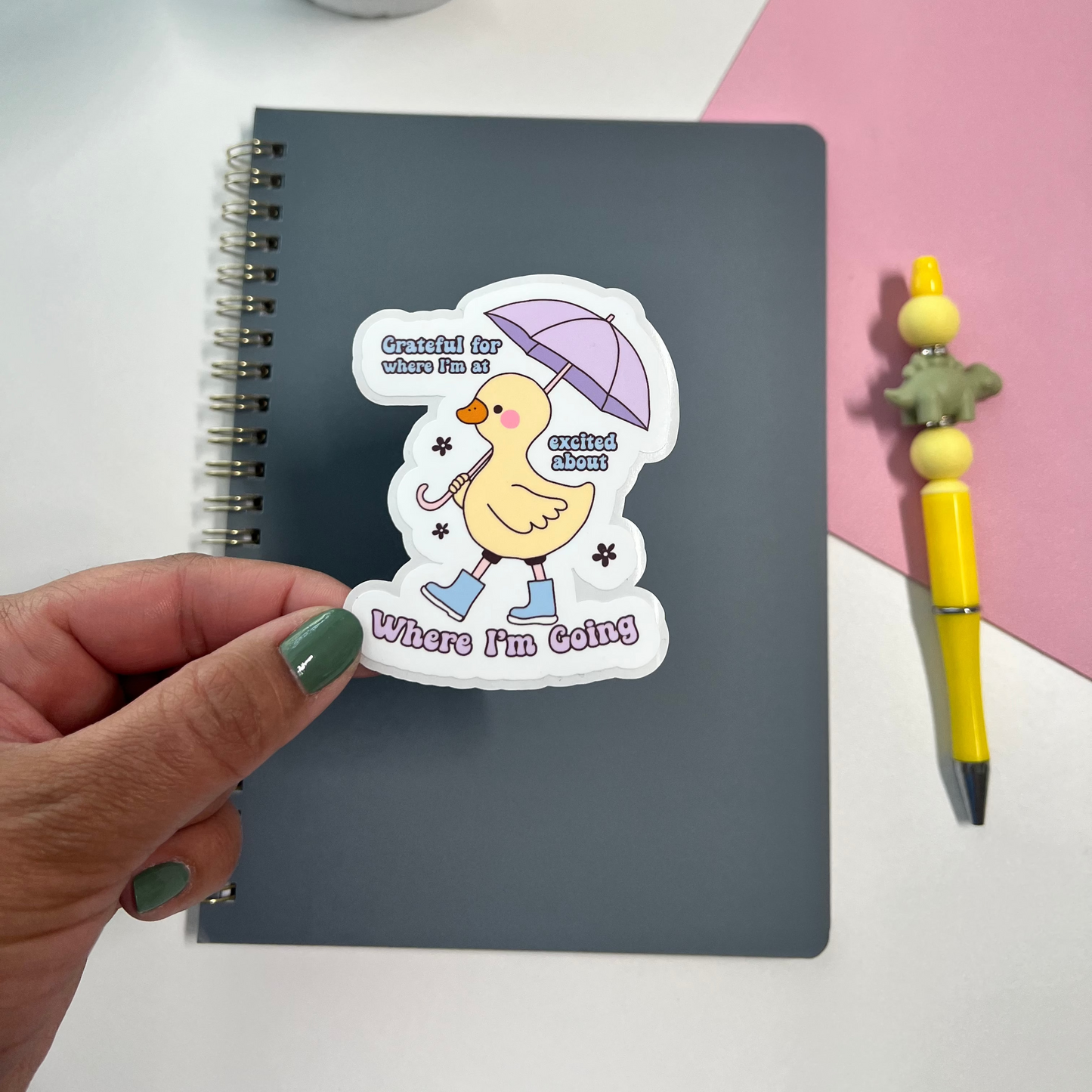 Sticker of a duck holding an umbrella with text held over a notebook on a white and pink background.