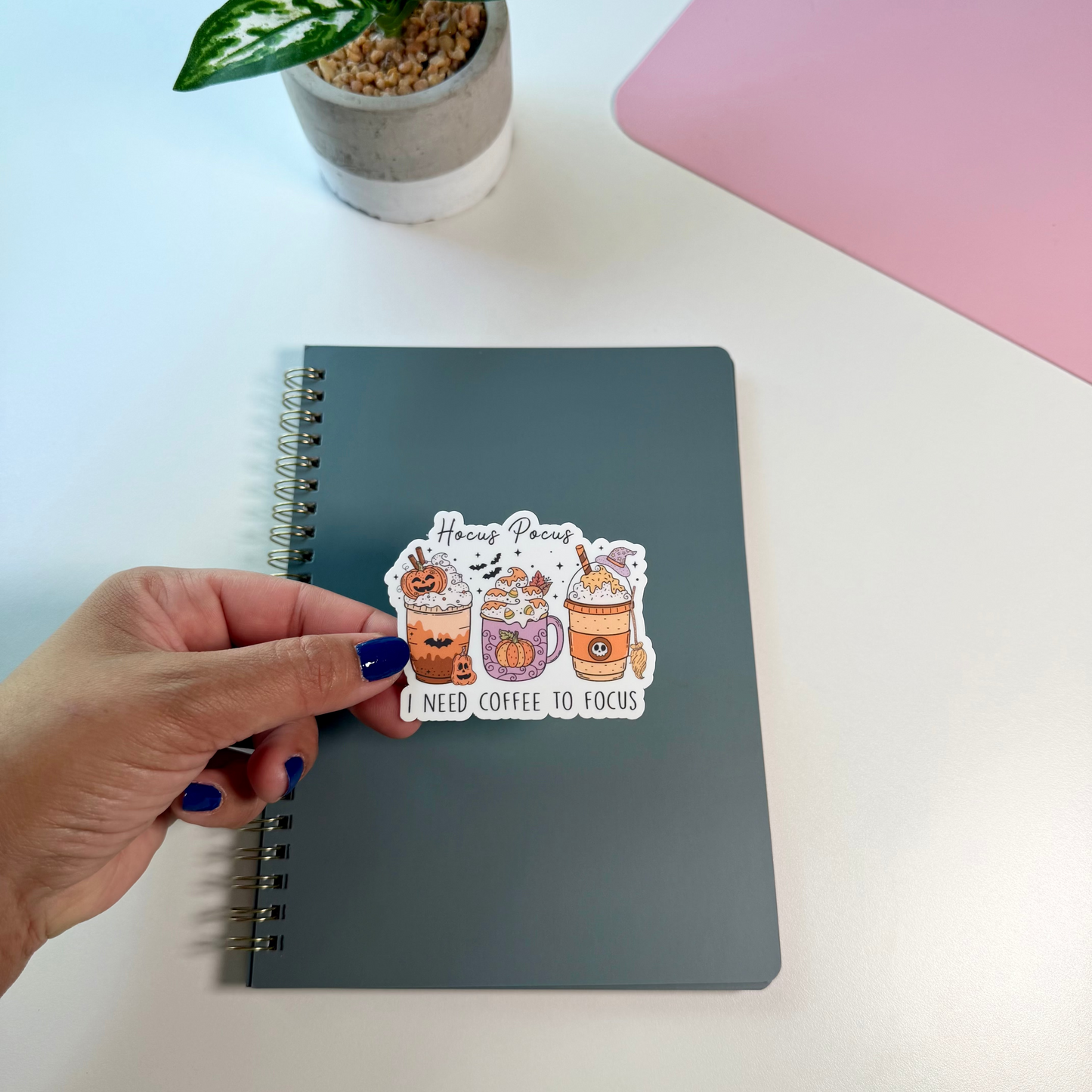 A hand with dark blue nail polish holds a die-cut sticker featuring three colorful coffee cups and the text 'Hocus Pocus I NEED COFFEE TO FOCUS' above a gray spiral notebook on a white desk. A potted plant and a pink laptop are visible in the background.