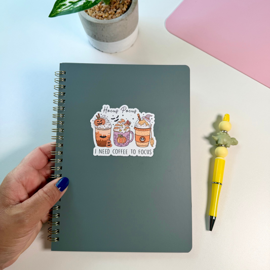 A dark gray spiral notebook with a cartoon sticker that says 'Hocus Pocus I Need Coffee to Focus' is held by a hand on a white desk, next to a yellow pen and a small potted plant.