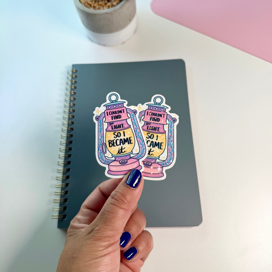 A hand with blue nail polish holds two decorative lantern stickers above a gray spiral notebook on a white desk, with a small potted plant and a pink mat visible in the background. 