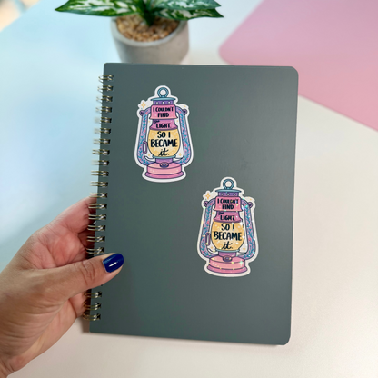 A hand with blue nail polish holds a dark gray spiral notebook decorated with two colorful lantern stickers that read 'I COULDN'T FIND THE LIGHT SO I BECAME IT'. The background shows a light-colored desk with a pink mat and a potted plant.