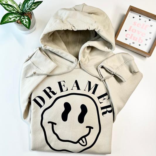 A beige pullover hoodie with a black graphic print featuring the word 'DREAMER' and a smiley face with its tongue sticking out, laid flat on a white surface with a small potted plant and a decorative frame nearby.
