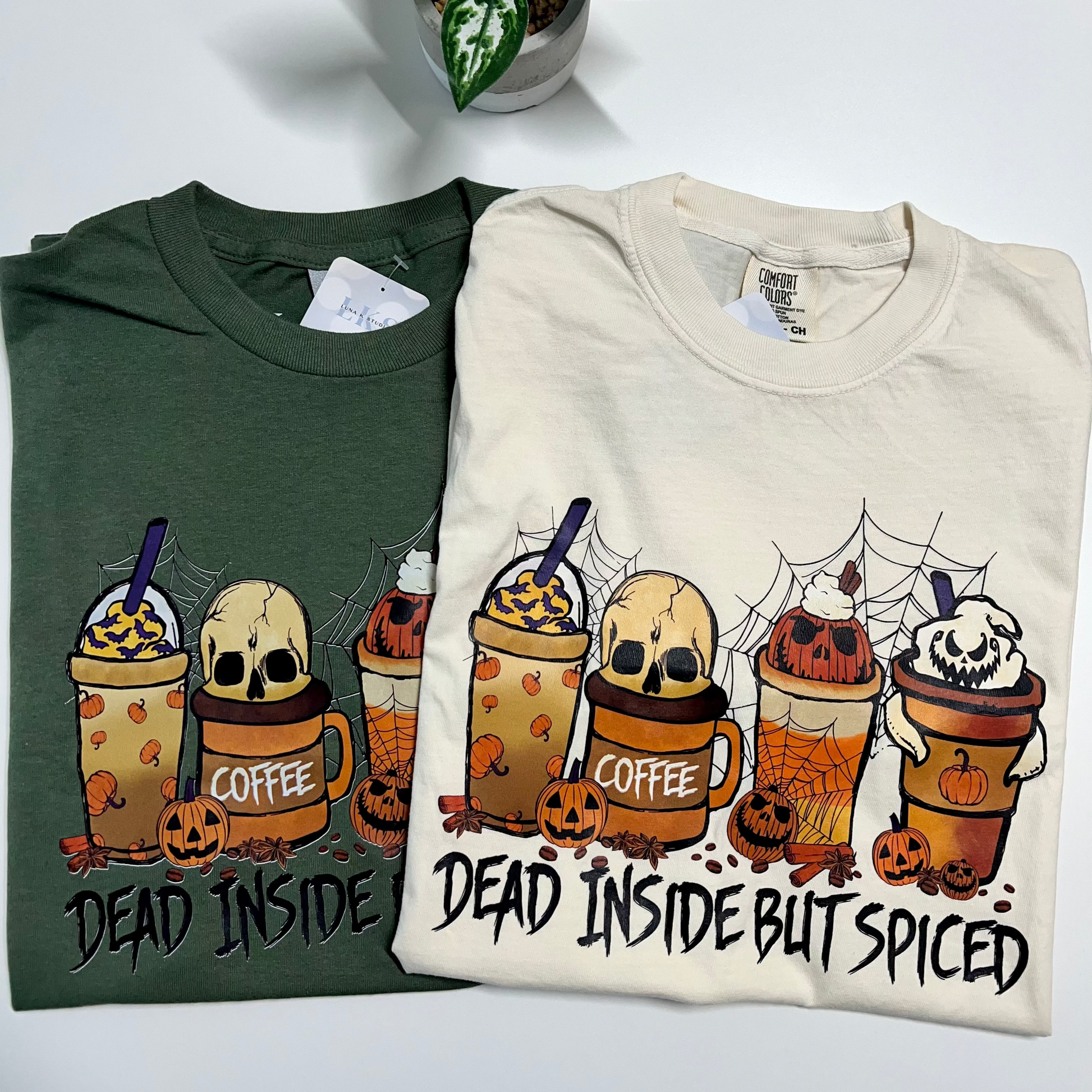 Two folded graphic T-shirts displayed side by side on a white surface with a small potted plant above. The left shirt is dark green and the right shirt is cream.