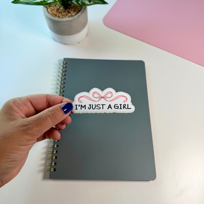 A hand with blue nail polish holds a white sticker that reads 'I'M JUST A GIRL' with a pink bow graphic, placed on a gray spiral notebook on a white desk. A small potted plant and a pink laptop are in the background.