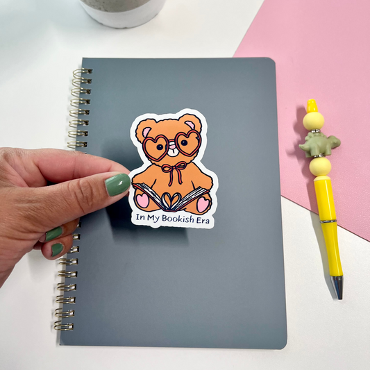 A hand with green nail polish holds a die-cut sticker featuring an orange teddy bear wearing glasses and holding a book, with the text 'In My Bookish Era' on a gray spiral notebook. The notebook is styled on a white desk with a pink paper background, a yellow pen, and a small potted plant.