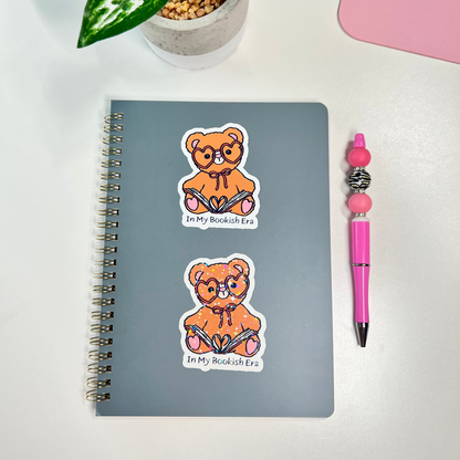A gray spiral-bound notebook with two cartoon teddy bear stickers on the cover, styled on a white surface with a potted plant, pink pen, and pink item in the background.