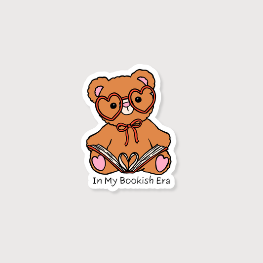 A die-cut sticker featuring a cartoon brown teddy bear wearing heart-shaped glasses and holding an open book, with the text 'In My Bookish Era' below it, shown on a light gray background.