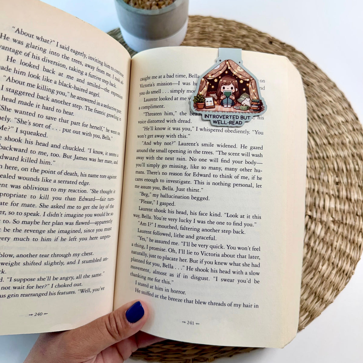 A hand with dark blue nail polish holds an open vintage book showing a printed illustration on the right page. A potted green plant is visible in the background on a white surface.