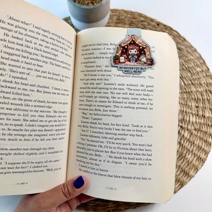 A hand with dark blue nail polish holds an open vintage book showing a printed illustration on the right page. A potted green plant is visible in the background on a white surface.