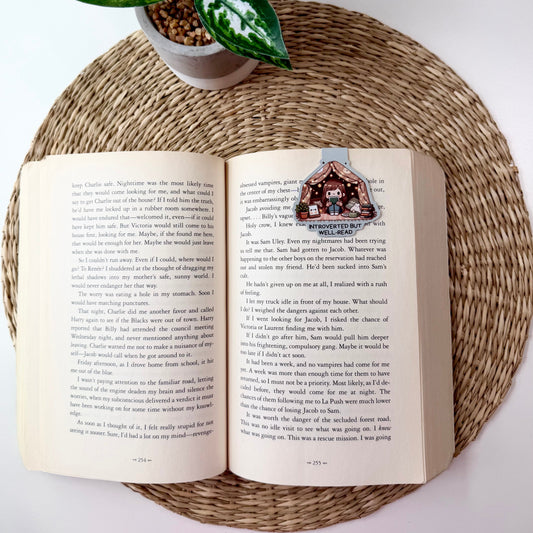 An open book spread with printed text and a small illustrated house at the top of the right page, styled on a round woven placemat with a potted green plant above it.