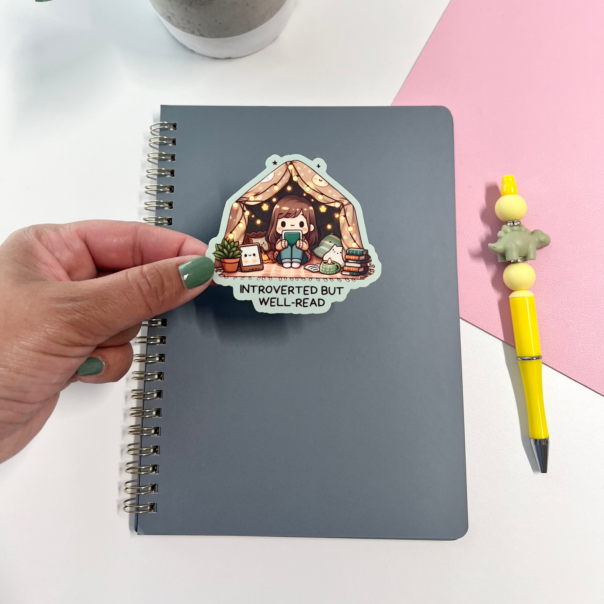 Notebook with a sticker of a cartoon character in a cozy room, held by a hand on a white surface with a plant and pen.