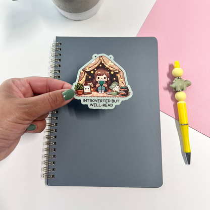 Notebook with a sticker of a cartoon character in a cozy room, held by a hand on a white surface with a plant and pen.