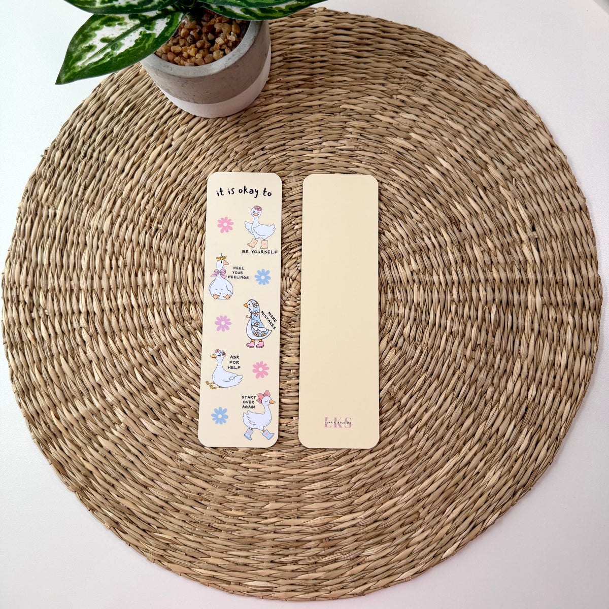 Two decorative paper bookmarks placed on a round woven placemat, with a small potted plant at the top left. One bookmark is cream with colorful cartoon illustrations and text, and the other is plain cream.