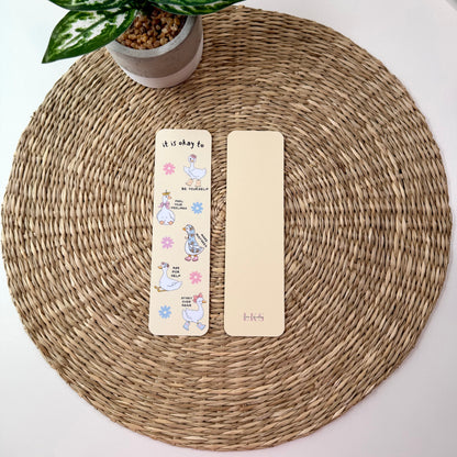 Two decorative paper bookmarks placed on a round woven placemat, with a small potted plant at the top left. One bookmark is cream with colorful cartoon illustrations and text, and the other is plain cream.