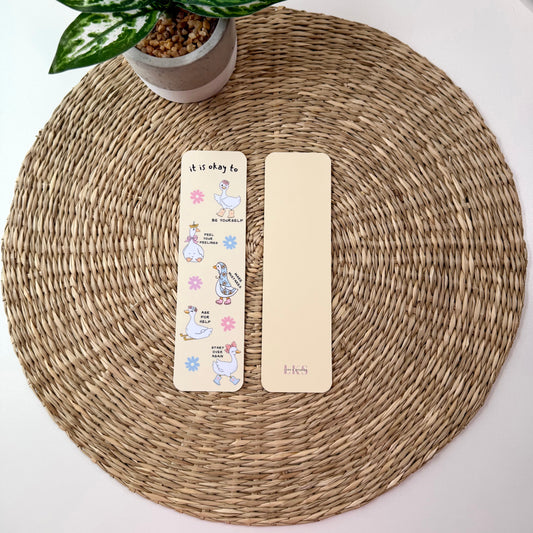Two decorative paper bookmarks placed on a round woven placemat, with a small potted plant at the top left. One bookmark is cream with colorful cartoon illustrations and text, and the other is plain cream.