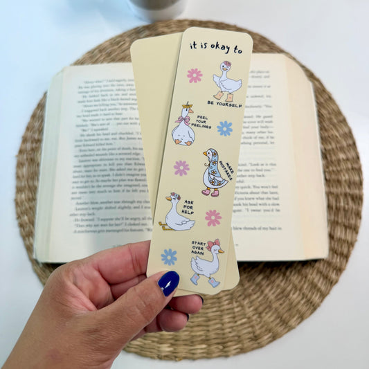 A hand with blue nail polish holds a cream-colored paper bookmark featuring cartoon ducks and the text 'it is okay to' over an open book on a woven placemat, with a small potted plant in the background.