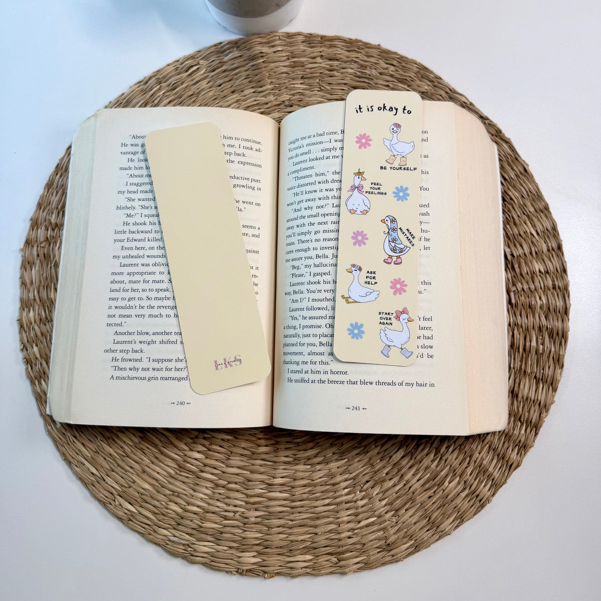 An open book resting on a round woven placemat, with a cream-colored bookmark or page flag on the left page and a decorative printed page with cartoon ducks on the right. A small potted plant sits above the setup on a white surface.
