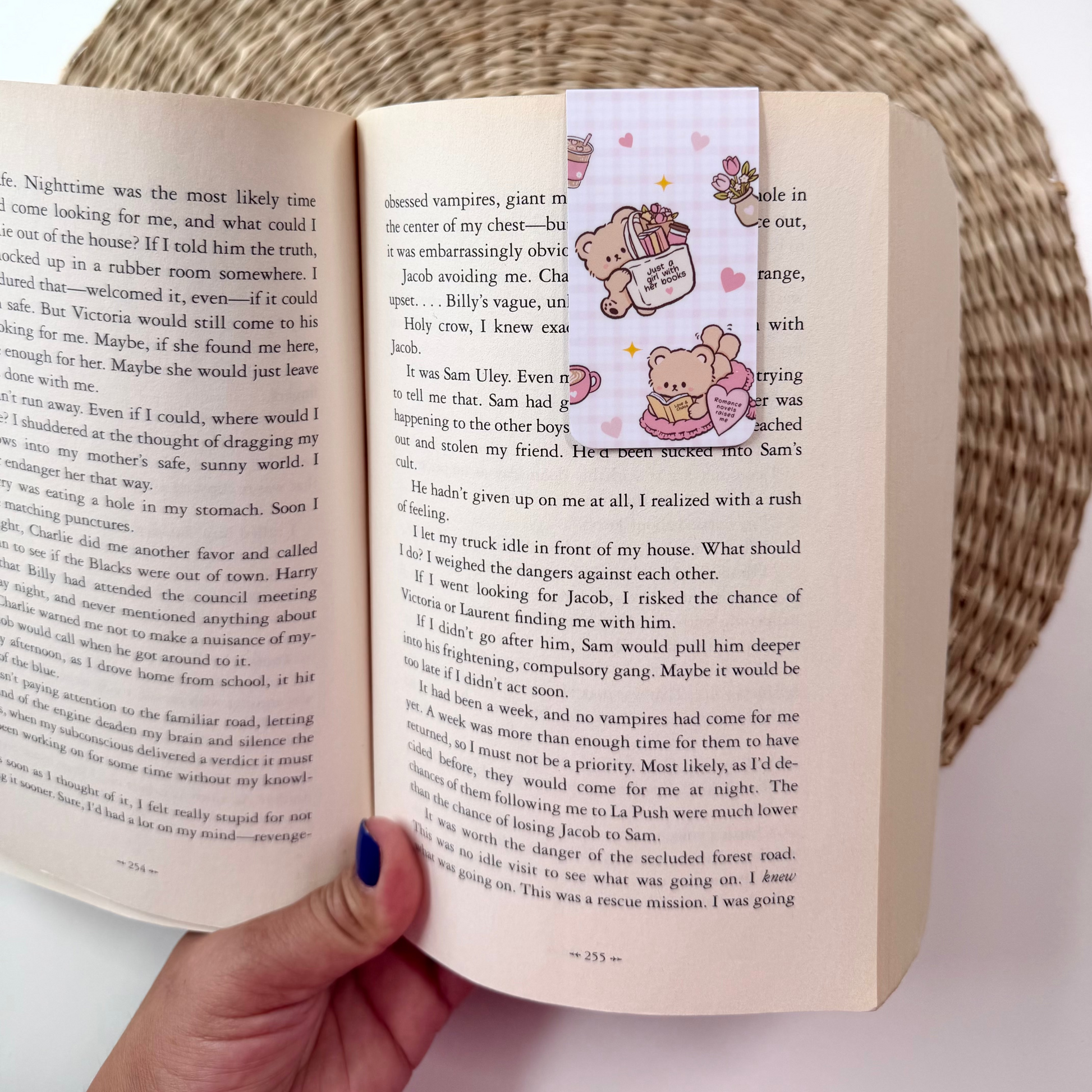 A hand holding an open book with printed text, featuring a decorative bookmark with cartoon teddy bears on the right page. The scene is styled with a woven placemat, a potted plant, and a bowl in the background.