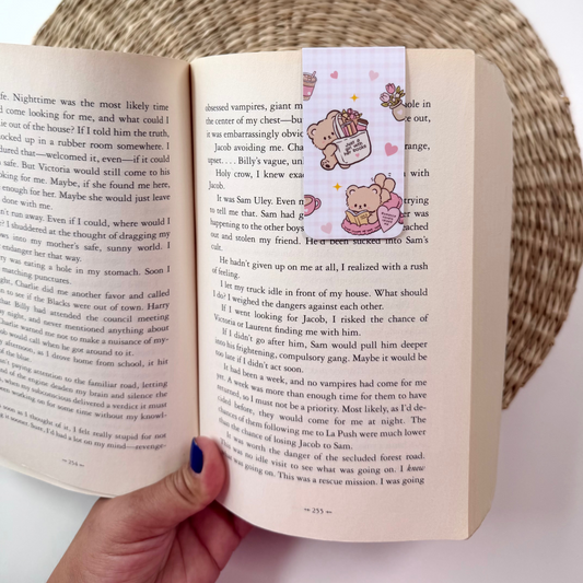 A hand holding an open book with printed text, featuring a decorative bookmark with cartoon teddy bears on the right page. The scene is styled with a woven placemat, a potted plant, and a bowl in the background.