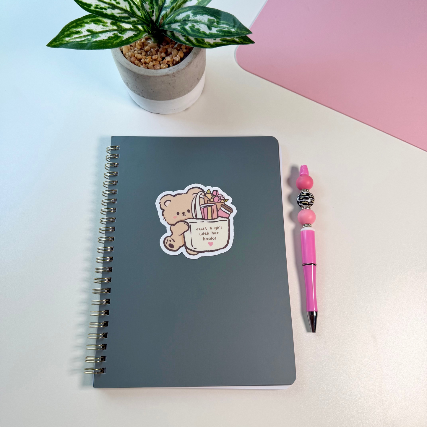 A dark gray spiral notebook with a cute cartoon bear sticker on the cover, styled on a white desk next to a pink pen and a small potted plant.