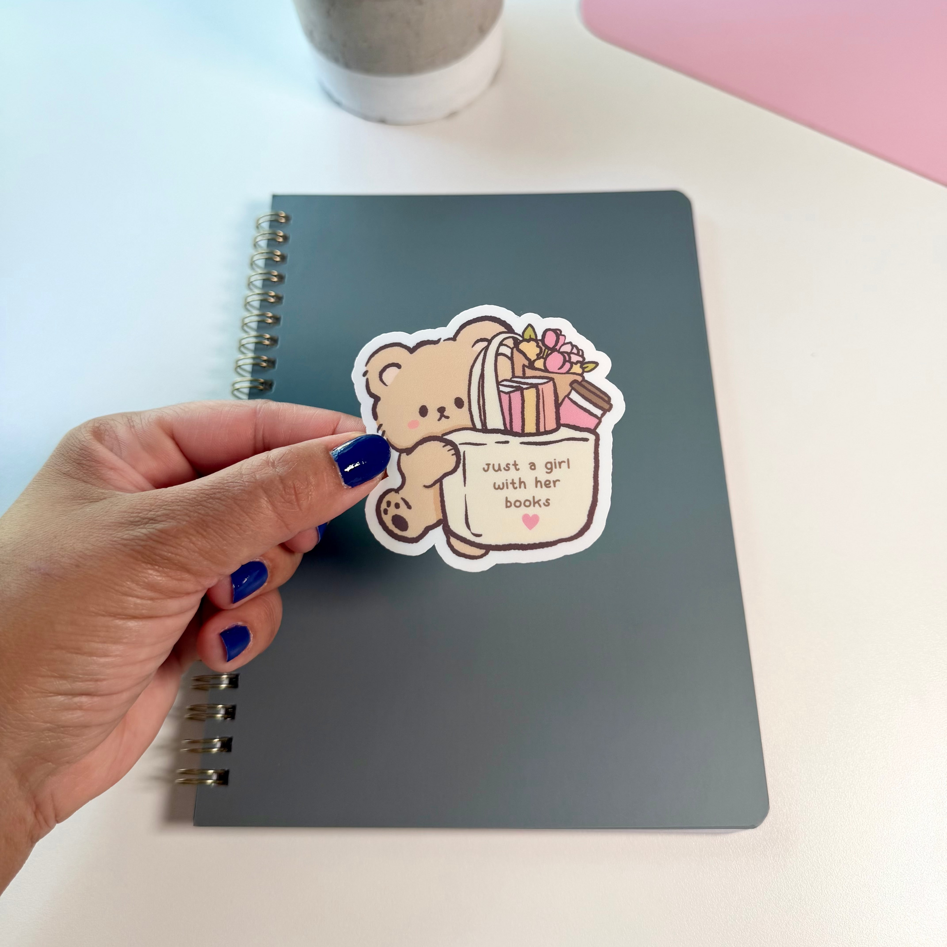 A hand with blue nail polish holds a die-cut sticker in front of a dark gray spiral notebook on a white desk. The sticker features a cute cartoon teddy bear holding a gift bag with the text 'want a girl with her books'.