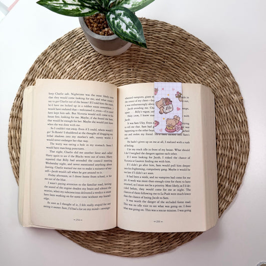 An open book with printed text and a small pink cartoon illustration on the right page, placed on a round woven placemat. A potted green plant sits at the top left, and part of a white desk or surface is visible around it.