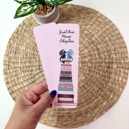 A hand with blue nail polish holds two light pink bookmarks featuring a book stack illustration and the text 'Just One More Chapter' on a woven placemat with a potted plant in the background.