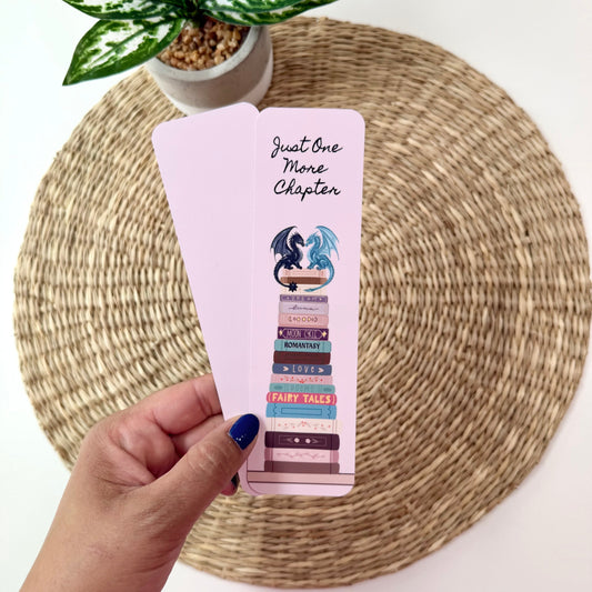 A hand with blue nail polish holds two light pink bookmarks featuring a book stack illustration and the text 'Just One More Chapter' on a woven placemat with a potted plant in the background.