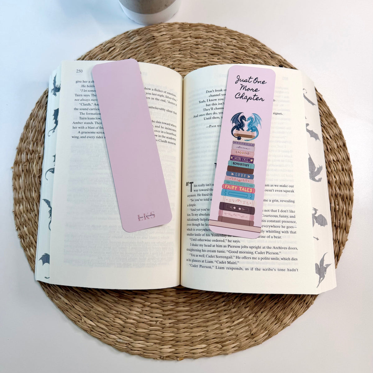 Two decorative bookmarks resting on an open book on a woven round placemat, with a small potted plant in the background. One bookmark is a solid pastel pink, and the other is a multicolor illustrated bookmark with a pink header reading 'Just One More Chapter'